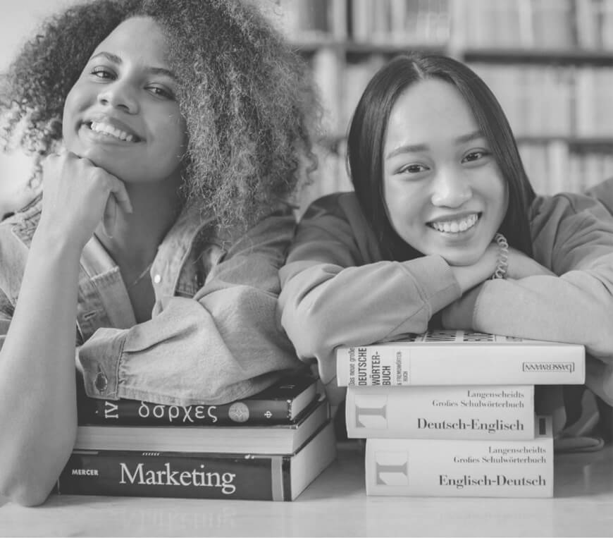 Two smiling young women leaning on stacks of books in a library.