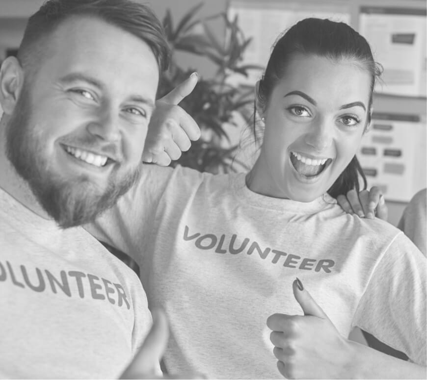 Smiling man and woman giving thumbs up while wearing volunteer t-shirts indoors.